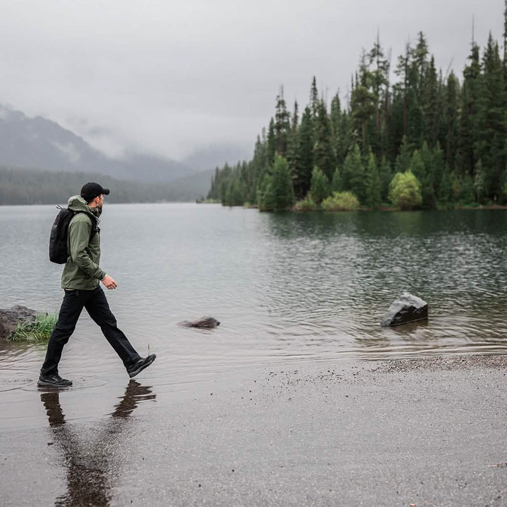 A person in a green jacket and black pants walks along a misty lakeshore, the dense evergreen trees framing their path. The overcast sky enhances the serene atmosphere, while their GORUCK Merino Challenge Socks - Trail Cuff ensure comfort with every step.