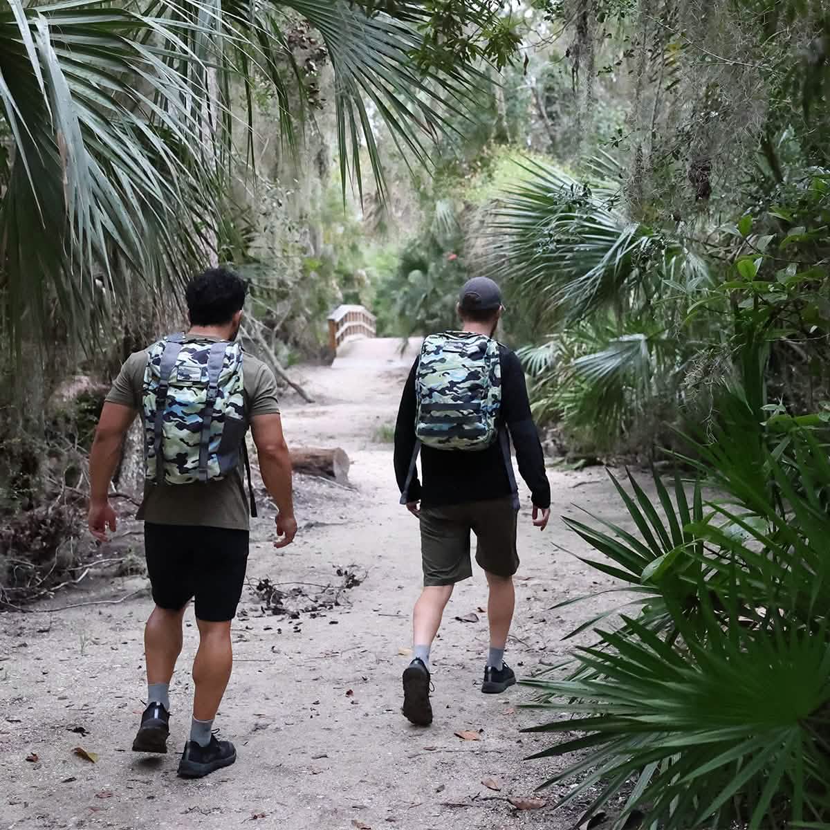 Two individuals equipped with GORUCK M24 - Cordura backpacks stroll along a path lined with vibrant greenery and gently swaying palm fronds. In the distance, a wooden bridge enhances the picturesque scene of their nature hike.