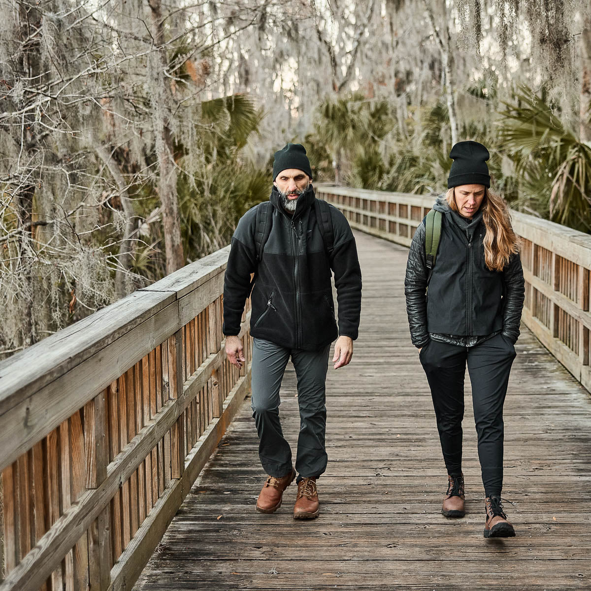 Two people in warm clothing, including Men’s Simple Pants - Midweight ToughDry®, walk on a wooden path through a forest with bare trees and hanging moss.
