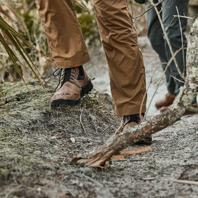Close-up of people wearing MACV-2 Mid Top shoes with an aggressive triple compound outsole and brown pants, walking on a dirt trail surrounded by foliage and fallen branches.