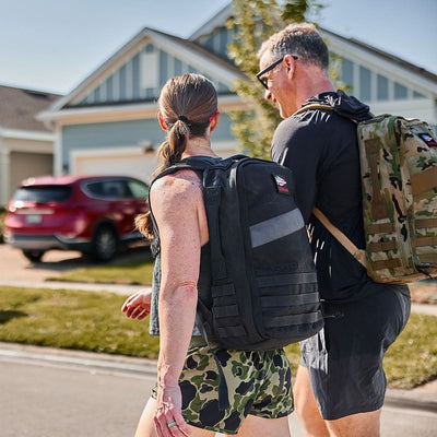 Two people with Rucker 4.0 rucksacks walk outside in a sunny suburban neighborhood.