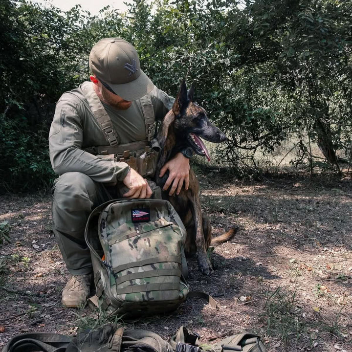 A man in military gear kneels beside a dog in a wooded outdoor area, carrying his GR1 USA - Ballistic Nylon Cordura (The Original Ruck) everyday carry rucksack.