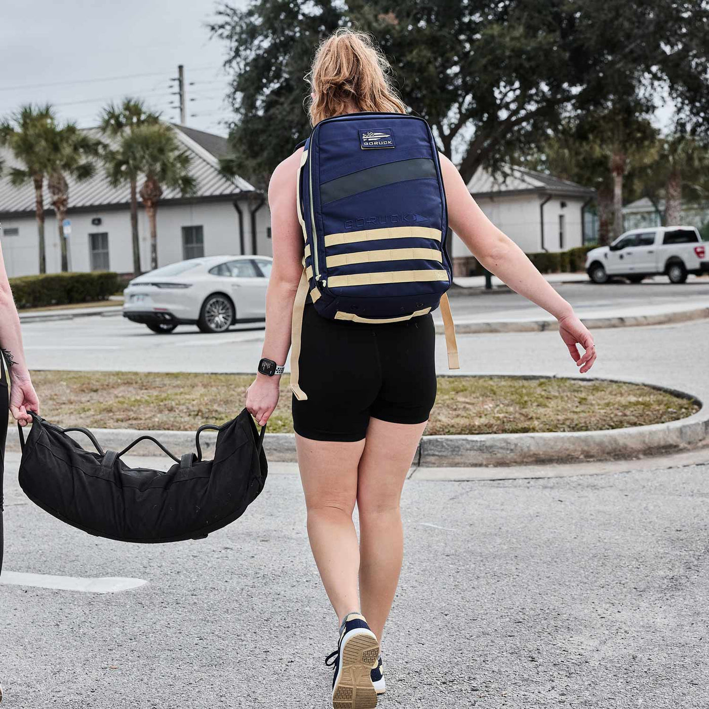 A person in athletic wear strides through the parking lot, carrying a GORUCK Rucker 4.0 loaded with ruck plates and a gym bag slung over their shoulder.