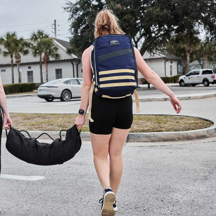 A person in athletic wear strides through the parking lot, carrying a GORUCK Rucker 4.0 loaded with ruck plates and a gym bag slung over their shoulder.
