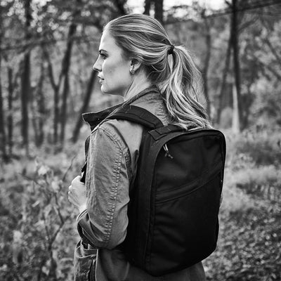 A woman with a ponytail stands in a forest, looking to the side and wearing the GR0 USA Slick backpack made from Ballistic Nylon Cordura. Black and white photo.