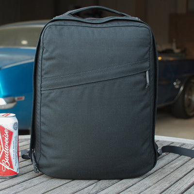 The GR0 USA Slick backpack in black Ballistic Nylon Cordura sits on a wooden table next to a Budweiser can, with a blue car visible in the background.