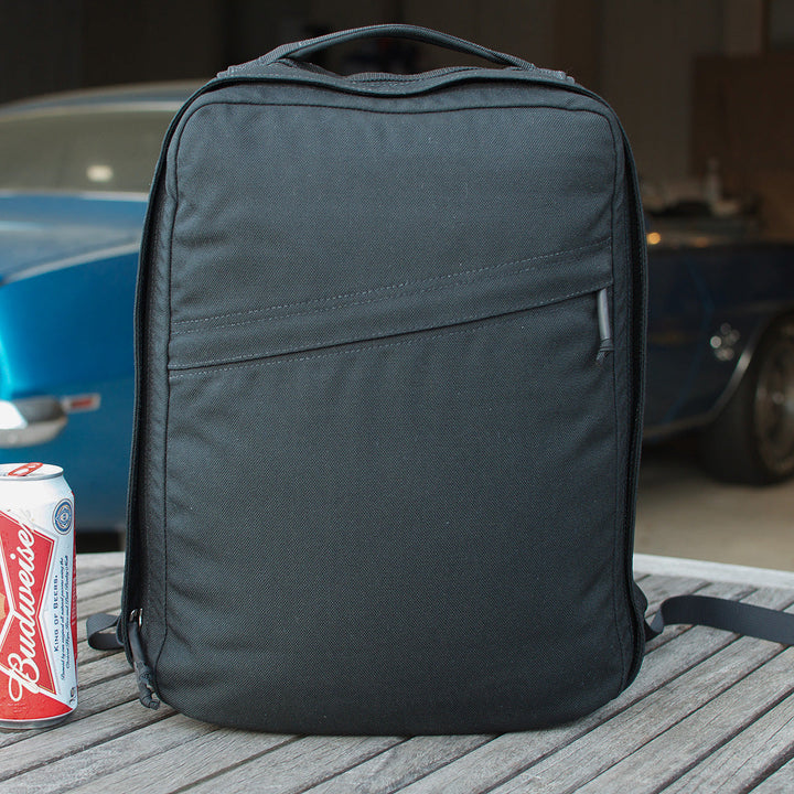 The GR0 USA Slick backpack in black Ballistic Nylon Cordura sits on a wooden table next to a Budweiser can, with a blue car visible in the background.