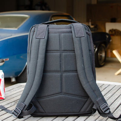 A black GR0 USA Slick - Ballistic Nylon Cordura backpack rests on a wooden table outdoors, with a blue vintage car in the background—a fusion of rugged style and Special Forces-level durability.