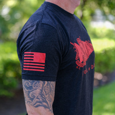 A man outdoors wears the Join or Die Tee - Tri-Blend, featuring a red flag and snake design from Ben Franklin—a tribute to colonial unity—while displaying his tattooed arm.
