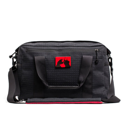 The Kit Bag x Carryology - GRXC2 Samurai 2.0 is a black duffel with handles, a shoulder strap, and a red patch with two black unicorns—crafted from custom technical fabric and inspired by the iconic Samurai Kit Bag design.