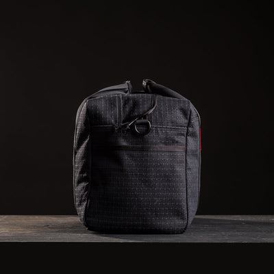 The Kit Bag x Carryology - GRXC2 Samurai 2.0 is a black, rectangular bag made from Japanese denim with a zipper and front pocket, shown against a dark background.