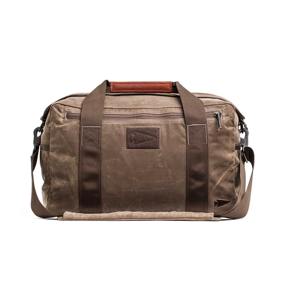 The Kit Bag - Waxed Canvas features brown waxed canvas with dark brown straps and a front zipper pocket. Ideal for travel, it combines durability and style to prepare you for any adventure.