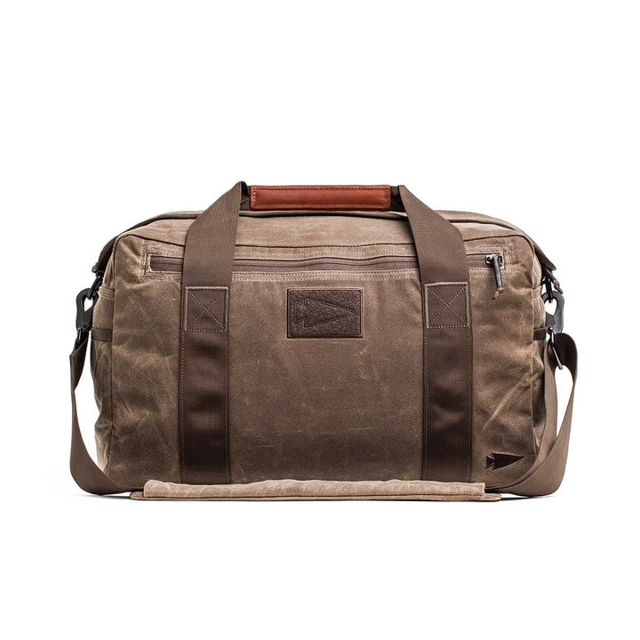 The Kit Bag - Waxed Canvas features brown waxed canvas with dark brown straps and a front zipper pocket. Ideal for travel, it combines durability and style to prepare you for any adventure.