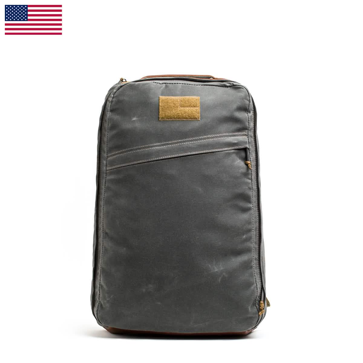 The GORUCK GR1 USA - Heritage rucksack, featuring a dark gray color with a front pocket and a small brown patch, is showcased against a plain white backdrop that highlights its minimalist design. An American flag adorns the top left corner, and it is expertly crafted from durable waxed canvas, making it both stylish and functional.