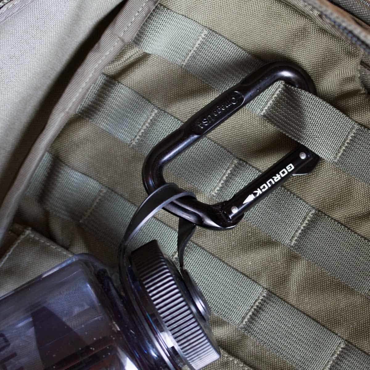 A clear water bottle is securely attached to a tactical backpack using a black climbing-certified SMC Gear Carabiner featuring the GORUCK Logo. The backpack boasts a green, grid-like strap design for secure attachment.