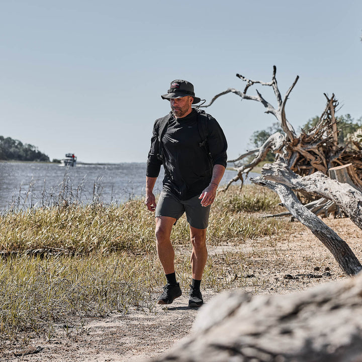 A man wearing the Men's USA Long Sleeve Performance Full Zip Hoodie - ToughMesh, a hat, and a backpack walks along a sandy riverside trail with driftwood and tall grass.