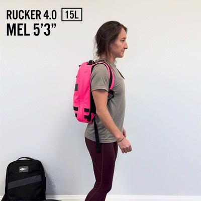 A woman wearing a pink backpack stands sideways, showcasing the Rucker 4.0, with "15L" and "Mel 5'3"" elegantly displayed, highlighting her rucking essentials.