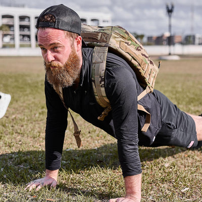 Man with a beard and camo backpack does a push-up on grass, wearing a black cap, outfit, and the Men's USA Long Sleeve Performance Half Zip - ToughMesh.