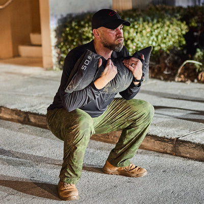 Man in camo pants and cap does a squat outdoors, holding a 50-lb sandbag while wearing the Men's USA Long Sleeve Performance Half Zip - ToughMesh, a featherweight training pullover made from durable ToughMesh™ fabric.