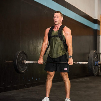 A man at the gym demonstrates his strength by lifting a barbell while wearing GORUCK’s Men’s USA Training Shorts made from ToughStretch fabric. Clad in a green tank top and white sneakers, with a fitness tracker on his wrist, he stands out against the gym's black and blue color scheme.