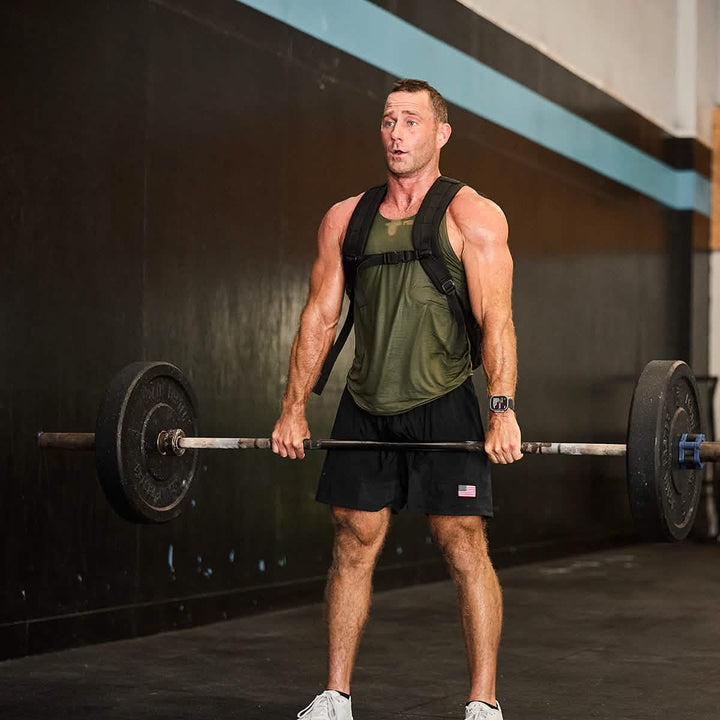 A man at the gym demonstrates his strength by lifting a barbell while wearing GORUCK’s Men’s USA Training Shorts made from ToughStretch fabric. Clad in a green tank top and white sneakers, with a fitness tracker on his wrist, he stands out against the gym's black and blue color scheme.
