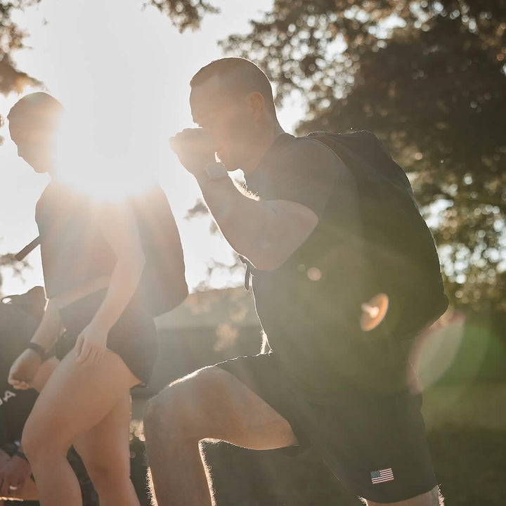 Two individuals exercise outdoors in bright sunlight, dressed in athletic clothing and backpacks. They're performing lunges in their GORUCK Men’s USA Training Shorts made from ToughStretch fabric. The sun creates a lens flare, partially obscuring their faces, with trees visible in the background.