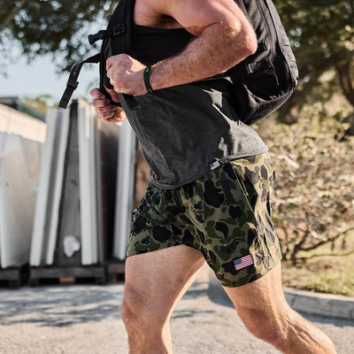 A man wearing GORUCK Men's USA Training Shorts - ToughStretch and a gray tank top jogs outdoors while carrying a black backpack. The shorts, featuring an American flag patch, are crafted from ToughStretch fabric for ultimate comfort and durability. In the background, trees and a stack of white panels can be seen.
