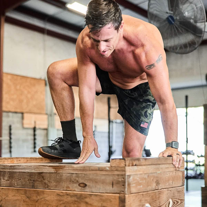 A man dressed in sports attire is executing a box jump in the gym, highlighting his Men’s USA Training Shorts from GORUCK, crafted with ToughStretch material. He is shirtless and wearing black sneakers. A large fan can be seen in the background, alongside various gym equipment, all offering resilience similar to the SCARS Lifetime Guarantee.