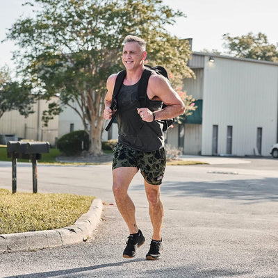A person jogging outdoors on a sunny day, sporting a tank top and GORUCK's Men’s USA Training Shorts crafted with ToughStretch fabric. They have a backpack slung over their shoulder and seem to be on a road near a building with trees in the background, epitomizing comfort and durability for every adventure.