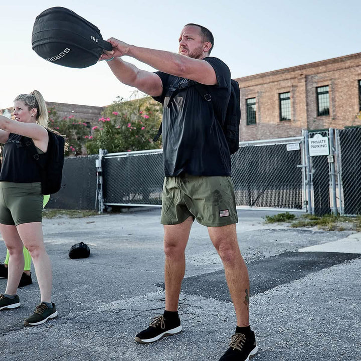 A man is engaged in an outdoor workout, gripping a weighted bag in front of him. He is wearing GORUCK's Men’s USA Training Shorts - ToughStretch and a black shirt, while also carrying a backpack. Nearby, another individual dressed in workout gear stands poised against the backdrop of a brick building and chain-link fence.