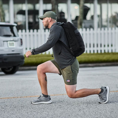 A man in GORUCK's Men’s USA Training Shorts - ToughStretch, a cap, and a long-sleeved shirt is briskly jogging along the street. With his black backpack bouncing, he passes by a parked car and white fence. His shorts are crafted with ToughStretch fabric for ultimate comfort on every run.