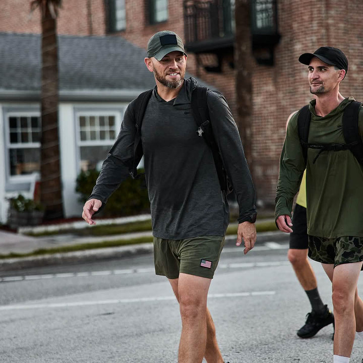 Two men in athletic apparel, demonstrating the resilience of GORUCK's Men’s USA Training Shorts - ToughStretch, stroll down a street carrying backpacks. They seem concentrated and deep in discussion. In the background, a brick building and a palm tree capture the essence of an ordinary day fueled by ToughStretch fabric performance.