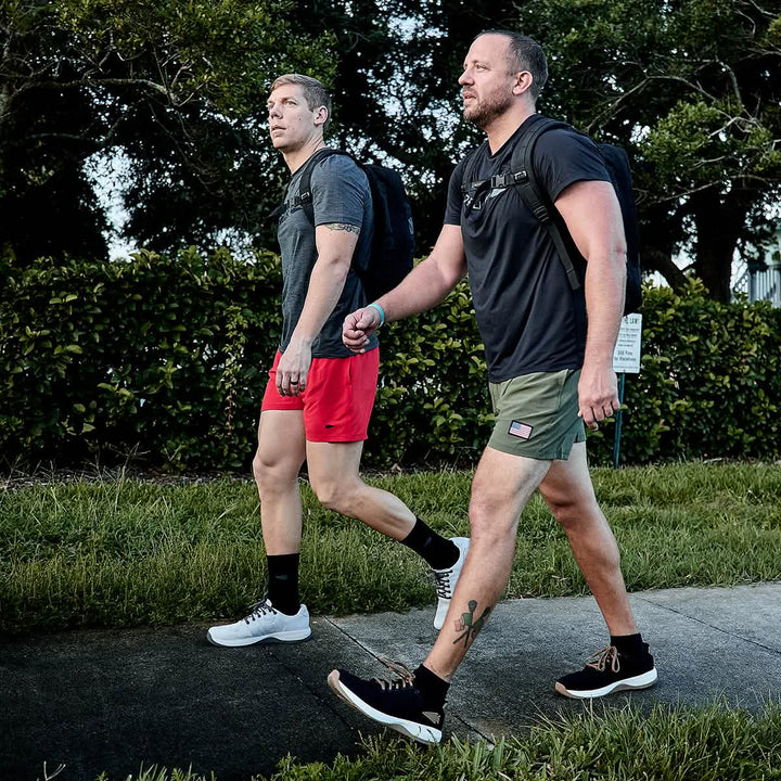 Two men walk along the sidewalk, sporting athletic gear and backpacks. The man in red shorts opts for Men’s USA Training Shorts by GORUCK, while his friend showcases his olive shorts featuring ToughStretch fabric, revealing a tattoo on his calf. Green bushes and trees frame their path as they stay focused and engaged in conversation.