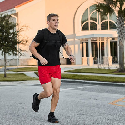 A man jogs down the street in Men's USA Training Shorts - ToughStretch by GORUCK, designed for ultimate comfort. His black t-shirt and backpack complete the ensemble, as a modern building with arched windows and palm trees provides a scenic backdrop in the warm climate.