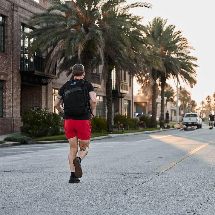 A person in a black shirt and Men’s USA Training Shorts by GORUCK jogs along a street lined with palm trees and brick buildings. The sun is low in the sky, casting long shadows. A few cars and cyclists are visible in the distance, enjoying the evening breeze.
