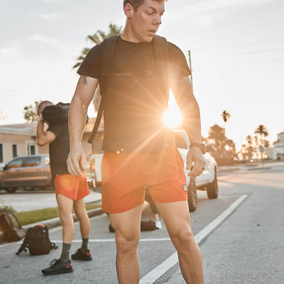 A person wearing a black shirt and GORUCK's Men’s USA Training Shorts - ToughStretch stands outdoors with a backpack, silhouetted by the sun. In the background, another individual adjusts a bag, while cars and palm trees line the street under a clear sky, highlighting the durability of the ToughStretch fabric in this sunny scene.