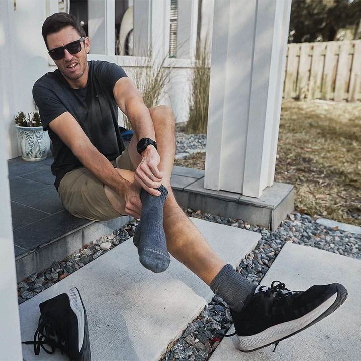 A man sitting on a porch step, wearing sunglasses and a watch, is putting on a gray Merino Challenge Sock from the GORUCK brand. His black shoes are placed on the ground next to him, and the area is surrounded by a garden with a white fence.