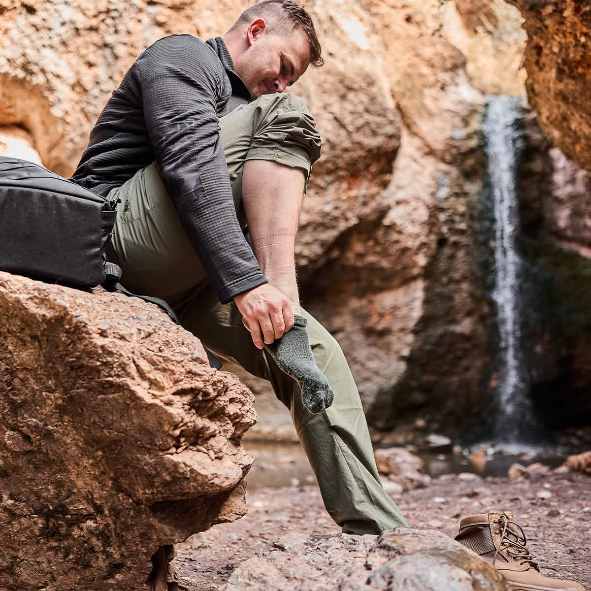 A person sits on a rock in a rocky landscape, adjusting their GORUCK Merino Challenge Socks - Crew. They wear a black jacket and khaki pants, with hiking boots placed beside them. A waterfall flows in the background, and a backpack rests nearby, showcasing the adventurous spirit of items Made in the USA.