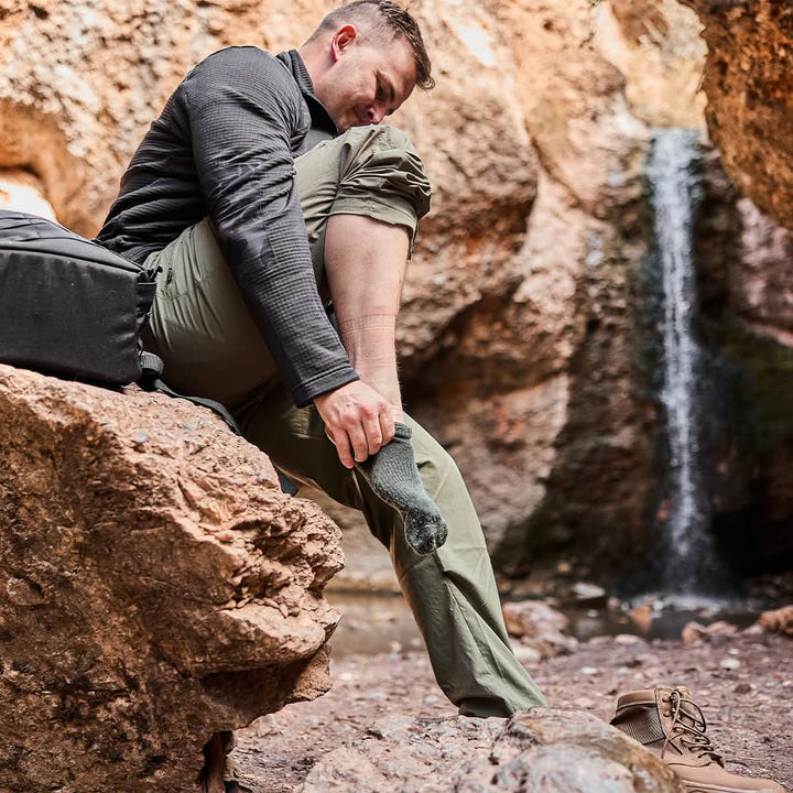 A person sits on a rock in a rocky landscape, adjusting their GORUCK Merino Challenge Socks - Crew. They wear a black jacket and khaki pants, with hiking boots placed beside them. A waterfall flows in the background, and a backpack rests nearby, showcasing the adventurous spirit of items Made in the USA.