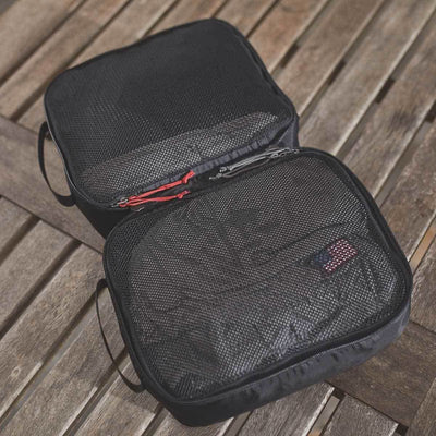 On a wooden surface lie two black GORUCK Packing Cubes, crafted to be ideal travel companions. Made from ultra-light nylon, one cube showcases red zipper details and a small flag label. They appear empty and ready to organize your travel essentials.