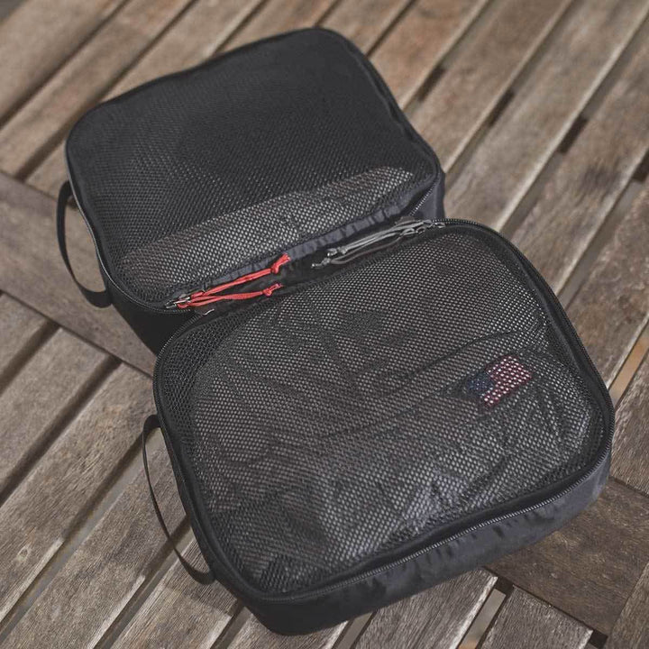 On a wooden surface lie two black GORUCK Packing Cubes, crafted to be ideal travel companions. Made from ultra-light nylon, one cube showcases red zipper details and a small flag label. They appear empty and ready to organize your travel essentials.