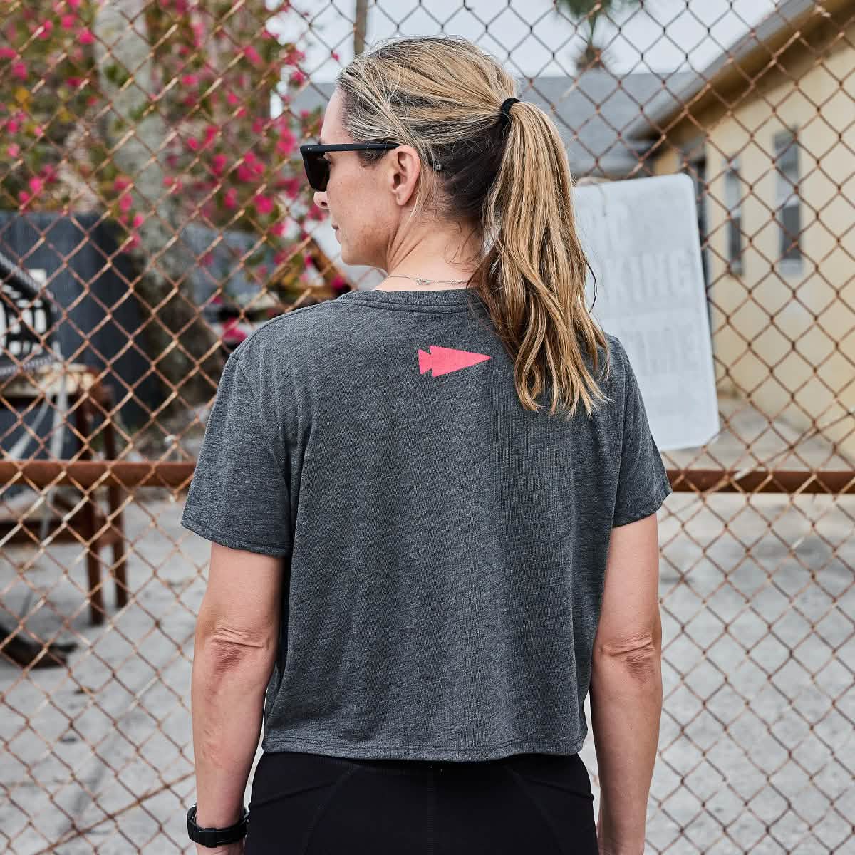 A woman with a ponytail stands outside near a chain-link fence, wearing a super soft Women's Cropped Tee from GORUCK, featuring a poly-blend fabric and a red arrow design on the back. In the background, flowers and a building suggest she's just finished her workout.