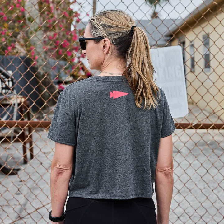 A woman with a ponytail stands outside near a chain-link fence, wearing a super soft Women's Cropped Tee from GORUCK, featuring a poly-blend fabric and a red arrow design on the back. In the background, flowers and a building suggest she's just finished her workout.