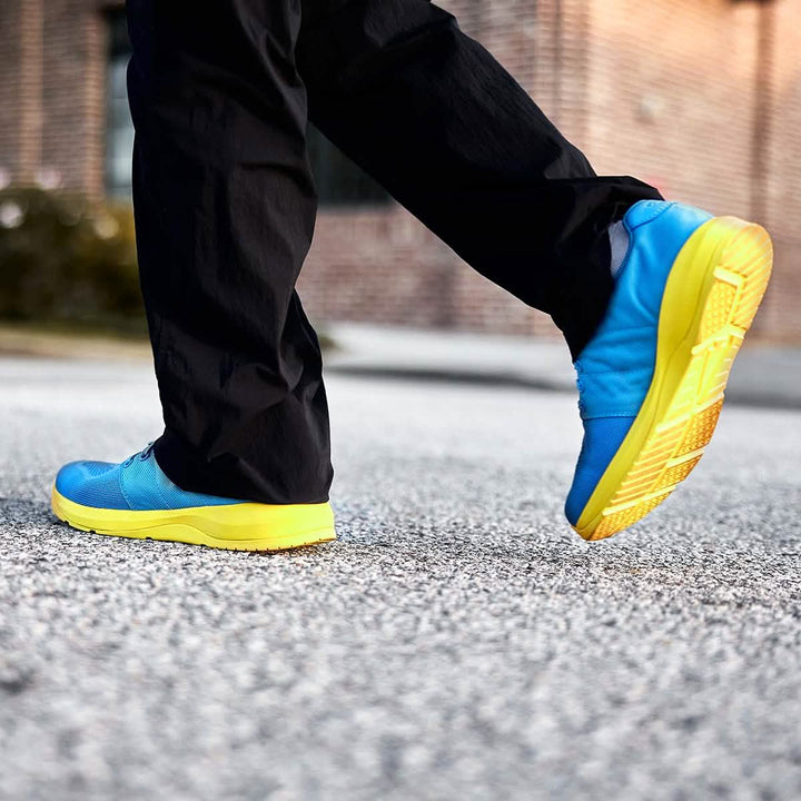 A person walks on pavement wearing Ballistic Trainers - Blue + Yellow, featuring 3X Support™ and yellow soles, paired with black pants.
