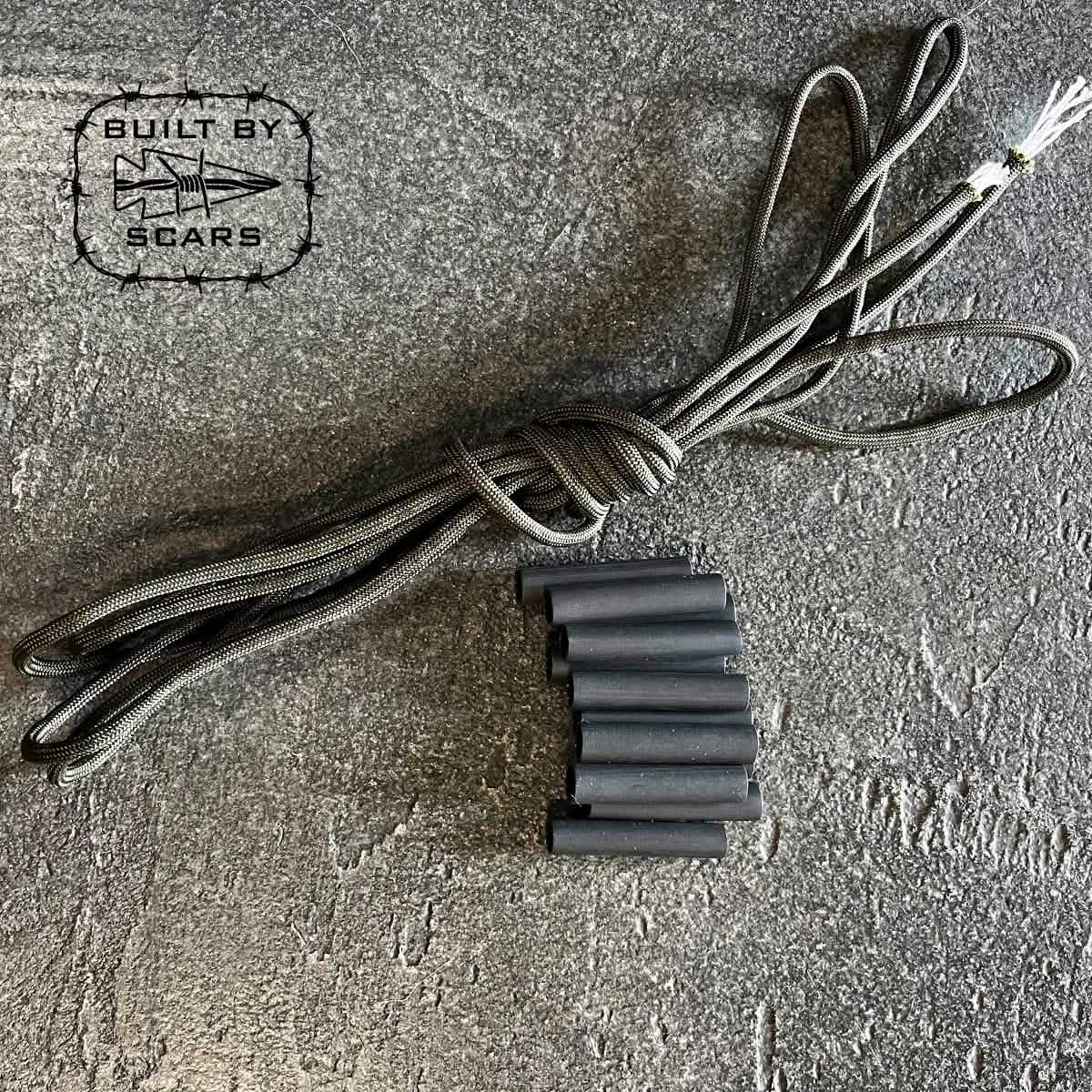 A coiled black rope, accompanied by multiple black tubes that appear to be heat shrink tubes, rests on a textured gray surface. In the top left corner, a logo showcasing a star and arrow displays the text "Paracord Kit - Built By SCARS" and highlights the GORUCK brand.