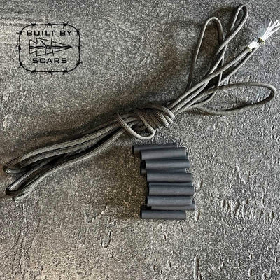 A coiled black rope, accompanied by multiple black tubes that appear to be heat shrink tubes, rests on a textured gray surface. In the top left corner, a logo showcasing a star and arrow displays the text "Paracord Kit - Built By SCARS" and highlights the GORUCK brand.