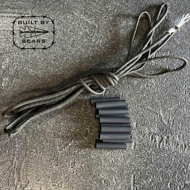 A coiled black rope, accompanied by multiple black tubes that appear to be heat shrink tubes, rests on a textured gray surface. In the top left corner, a logo showcasing a star and arrow displays the text "Paracord Kit - Built By SCARS" and highlights the GORUCK brand.