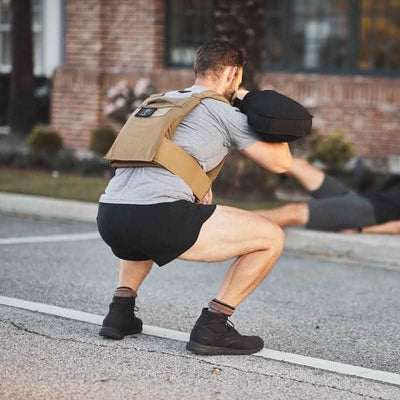A person wearing a weighted vest and Men’s Ranger Panties - ToughStretch squats while holding a sandbag outdoors. In the background, another person lies on the ground. They are on a paved surface with a brick building and plants visible, demonstrating the durability guaranteed by GORUCK's Scars Lifetime Guarantee.
