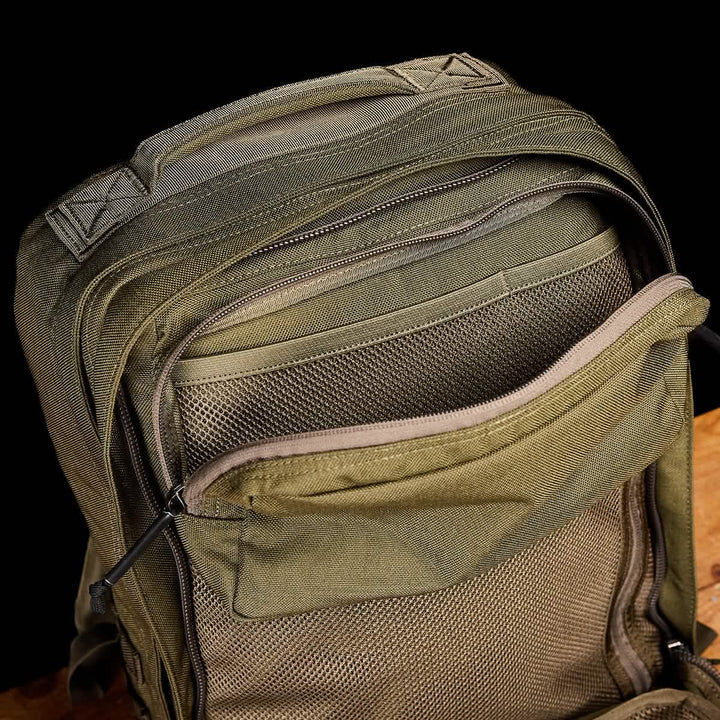 A close-up of a green GR2 - Cordura by GORUCK highlights its numerous zippered compartments. The ruck is open, displaying mesh-lined pockets, set against a dark background on a wooden surface, evoking an aura of Special Forces efficiency.
