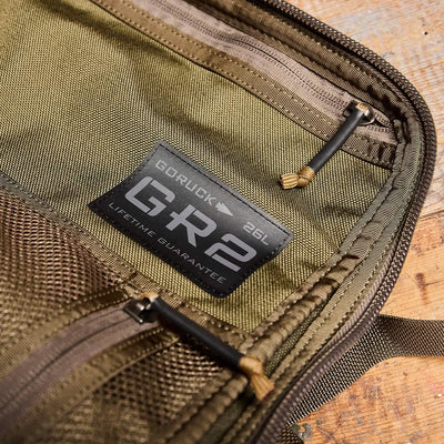 Close-up of an olive green rucksack labeled GORUCK GR2 - Cordura, featuring a Lifetime Guarantee. The image showcases the zipper details and stitching, emphasizing its durability and quality inspired by Special Forces. The bag is partially unzipped, revealing its interior.